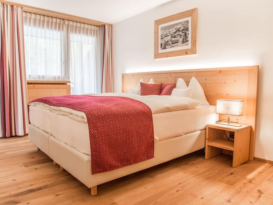 Ein Schlafzimmer mit einem hölzernen Kopfteil, einem Bett mit roter Decke, einem Nachttisch mit Lampe und einem Fenster mit Vorhängen.