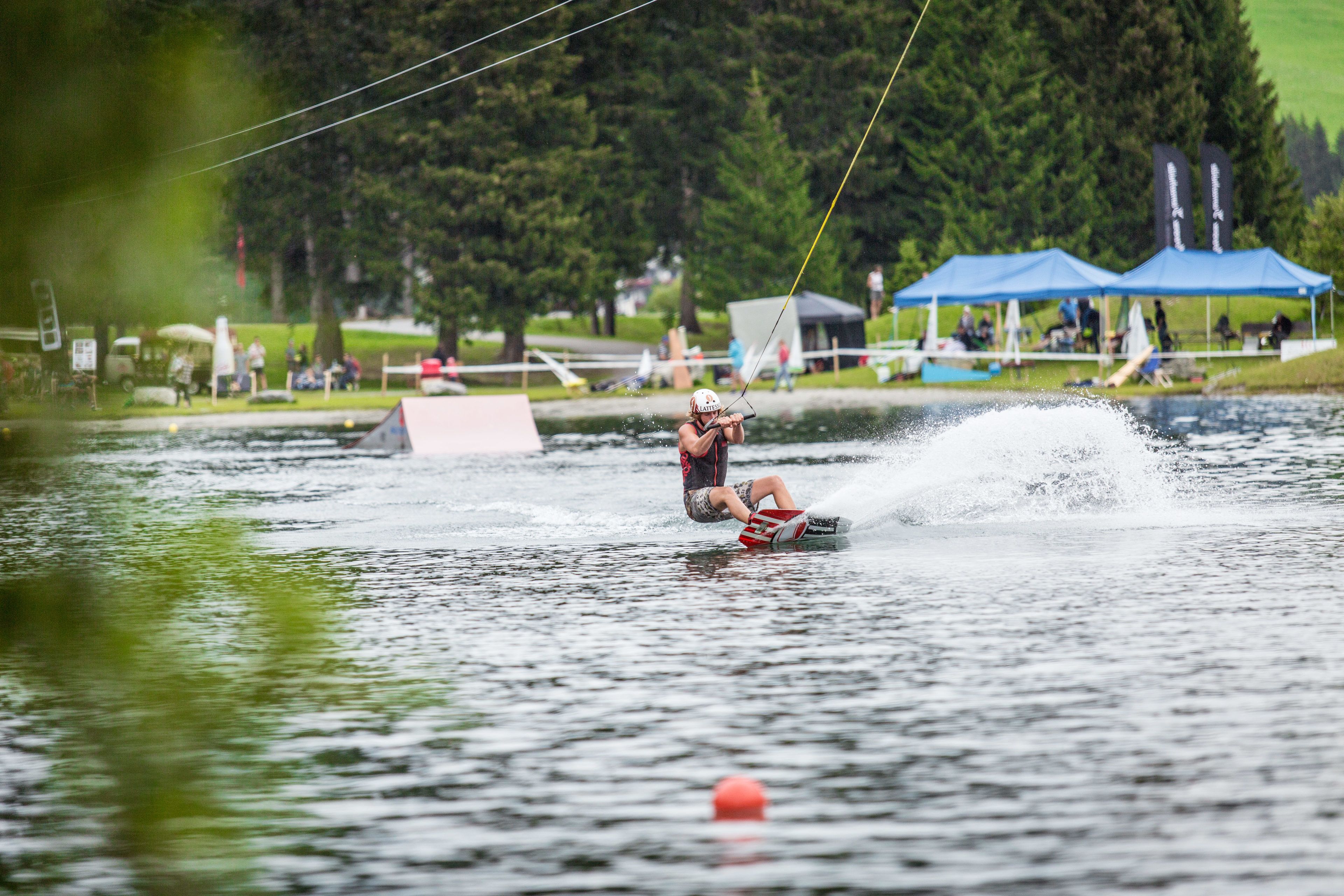 Evento de wakeboard com um praticante de wakeboard em pleno movimento em um lago, cercado por barracas e espectadores.
