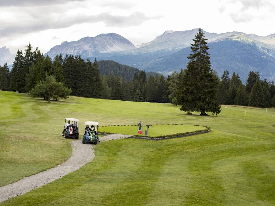 A golf course with a path leading to a tee box, surrounded by trees and mountains in the background.