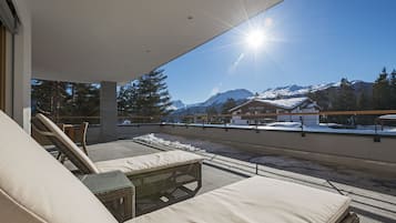 Eine Dachterrasse mit Liegestühlen und einem Korbtisch, mit Blick auf eine verschneite Berglandschaft und ein Gebäude mit der Aufschrift 'Hotel Barach'.
