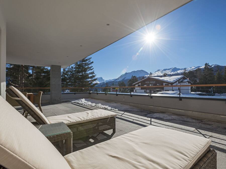 A rooftop terrace with lounge chairs and a wicker table, overlooking a snowy mountain landscape and a building labeled "Hotel Barach."