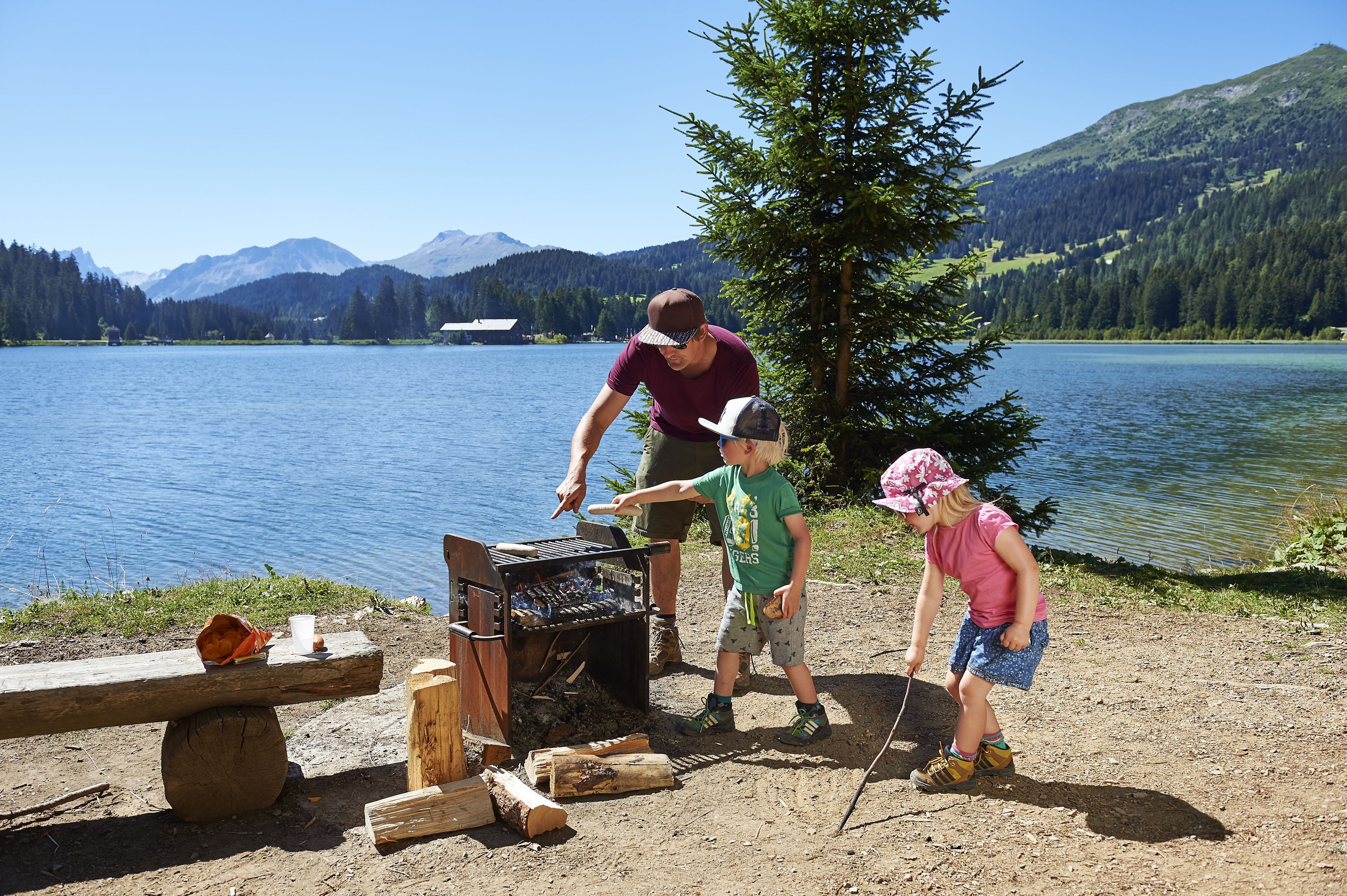 A lakeside setting with a barbecue grill, a picnic table, and a forested mountain backdrop.