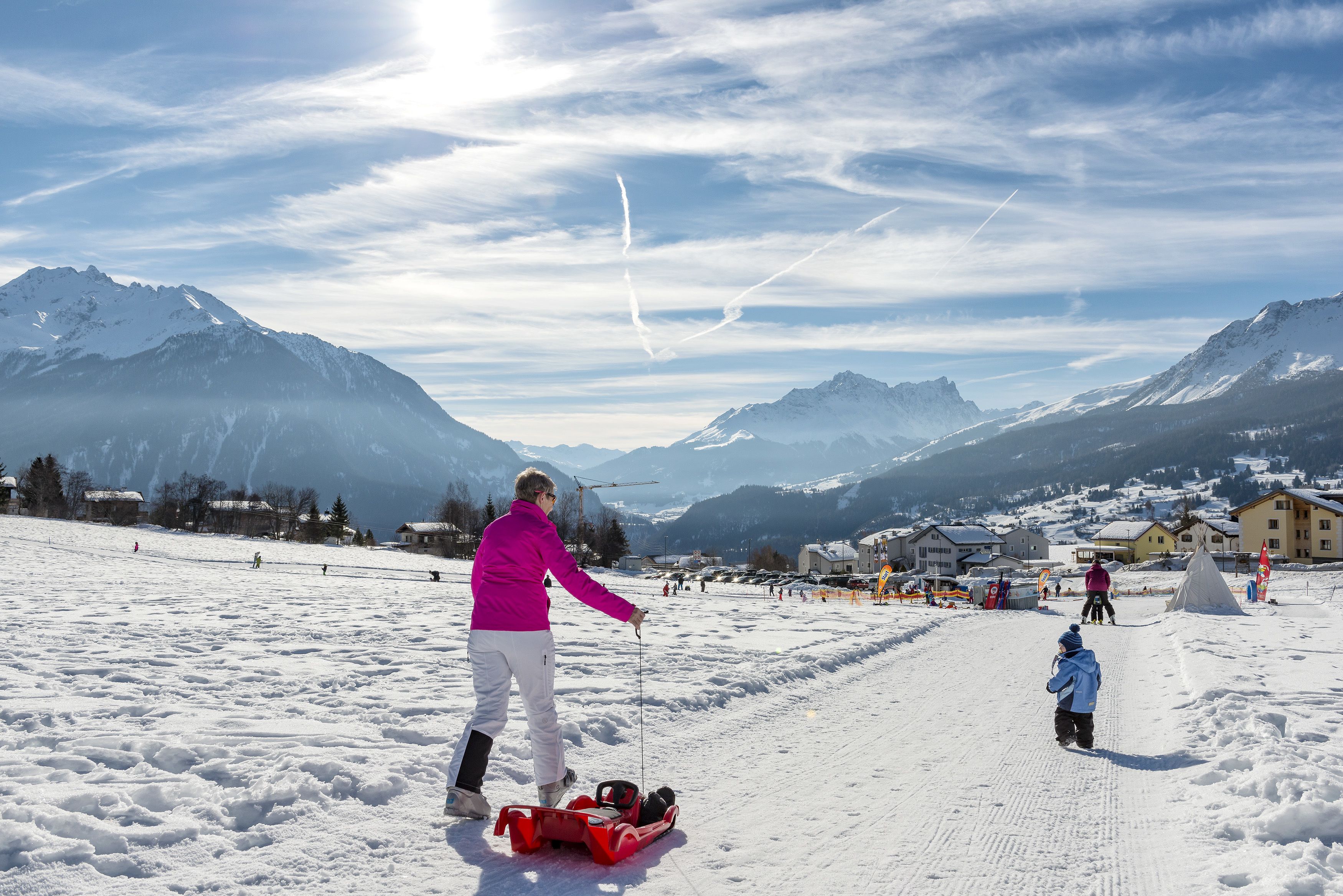 A snowy ski slope with a person in a pink jacket and white pants, a child in a blue jacket, and a red sled.