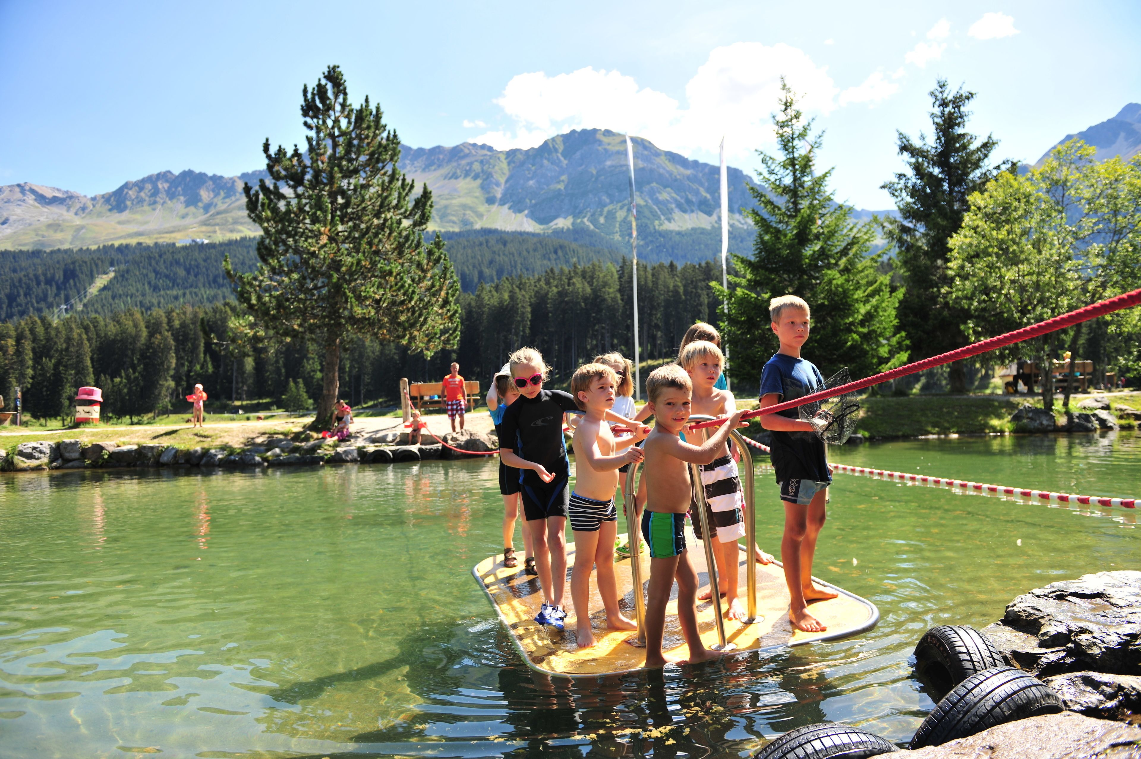 A group of people standing on a floating platform in a lake with mountains in the background.