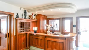 A reception area with a wooden reception desk, a large vase with flowers, and a decorative shelf with figurines. - Hotel Bellaria (Levico Terme)