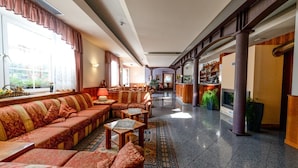 A hotel lobby with a long sofa, armchairs, a bar area, and a reception desk. - Hotel Bellaria (Levico Terme)