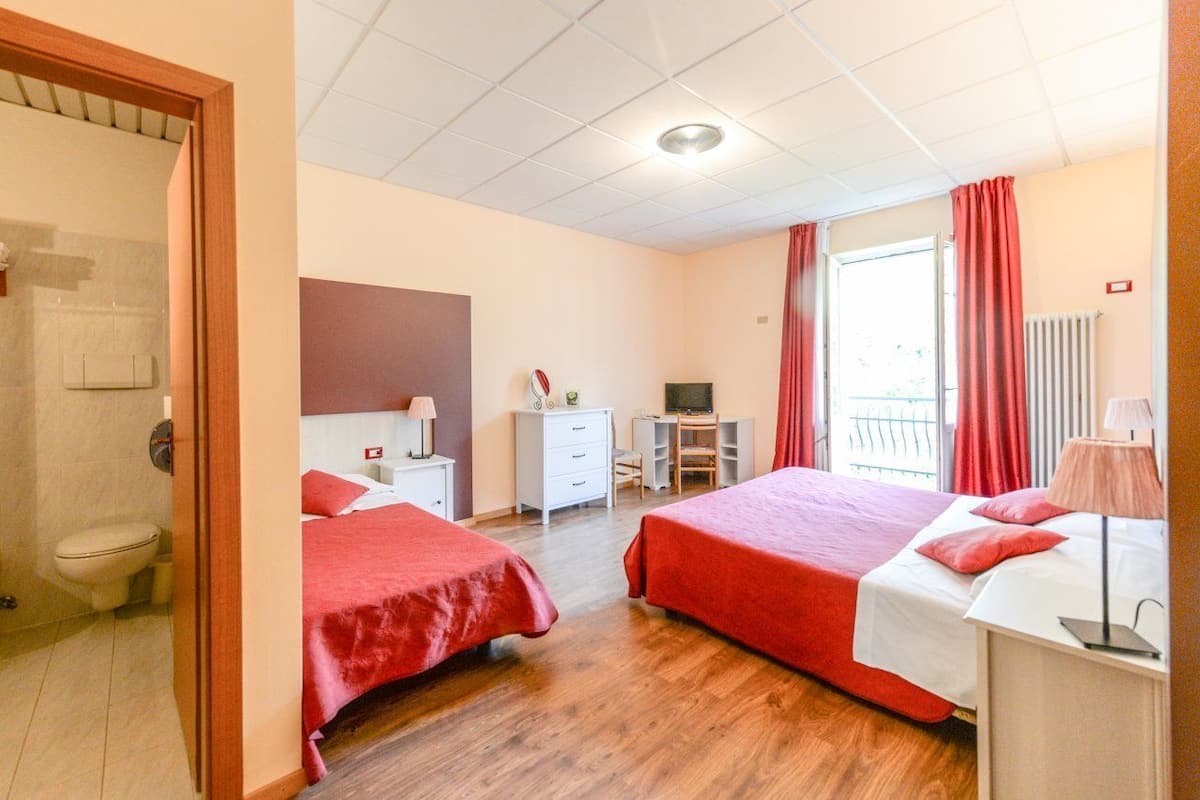 A hotel room with two beds, a desk, a chair, a small table, a radiator, and a window with curtains.