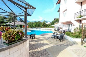 Piscina com espreguiçadeiras e guarda-sóis, rodeada por um edifício com varandas e um jardim.