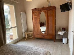 Quarto com um guarda-roupa de madeira grande, uma mesinha com um vaso, um radiador e uma televisão fixada na parede.