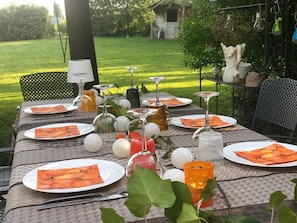Mesa de jantar ao ar livre posta com pratos, talheres e copos, rodeada por vegetação e uma pequena construção ao fundo.