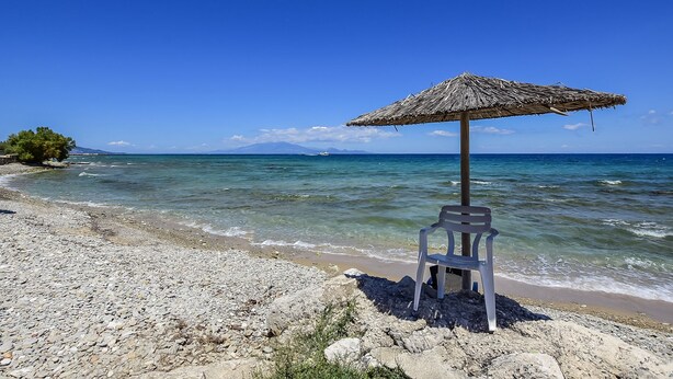 A beach with a thatched umbrella and a plastic chair, clear blue sea, rocky shore, and distant mountains. - Porto Giardino Apartments (Zakynthos)