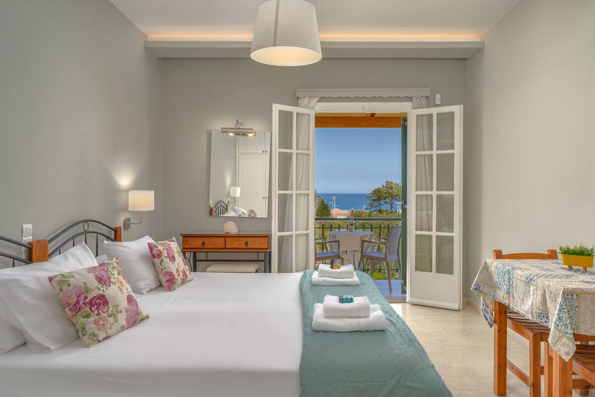 A bedroom with a bed, a dresser, a mirror, and an open door leading to a balcony with a table and chairs.