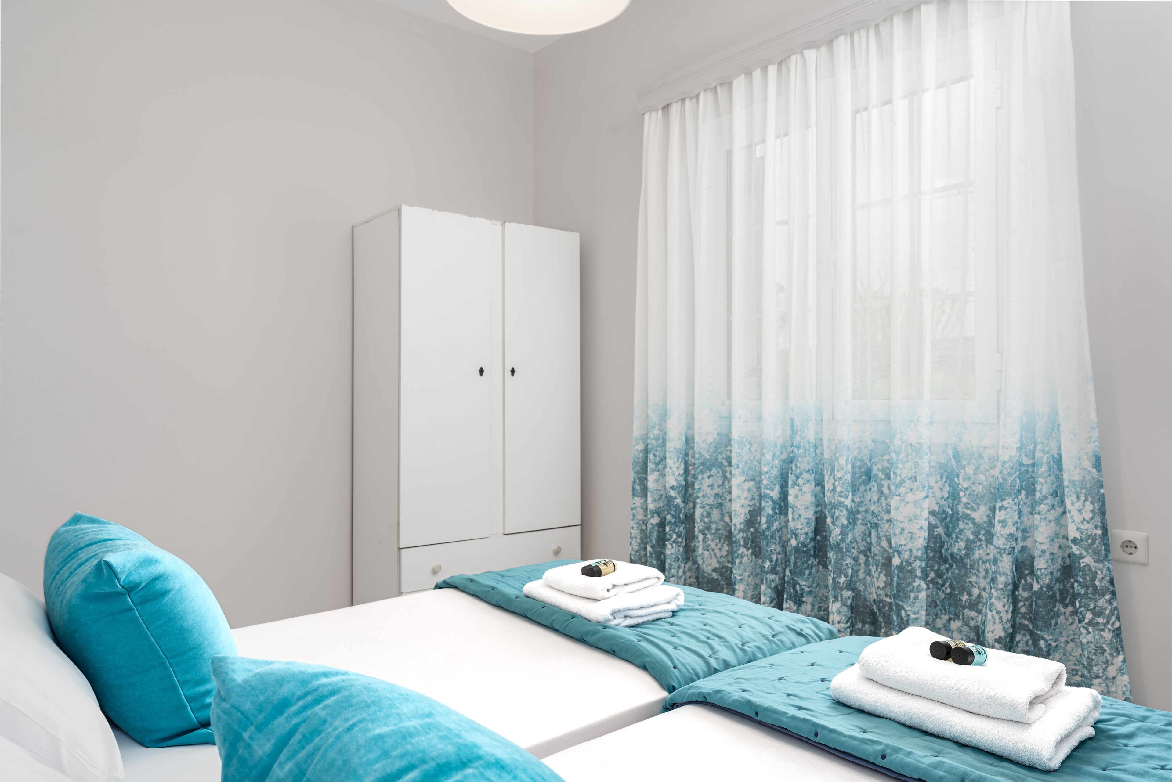 A bedroom with two beds, a wardrobe, and a window with sheer curtains.