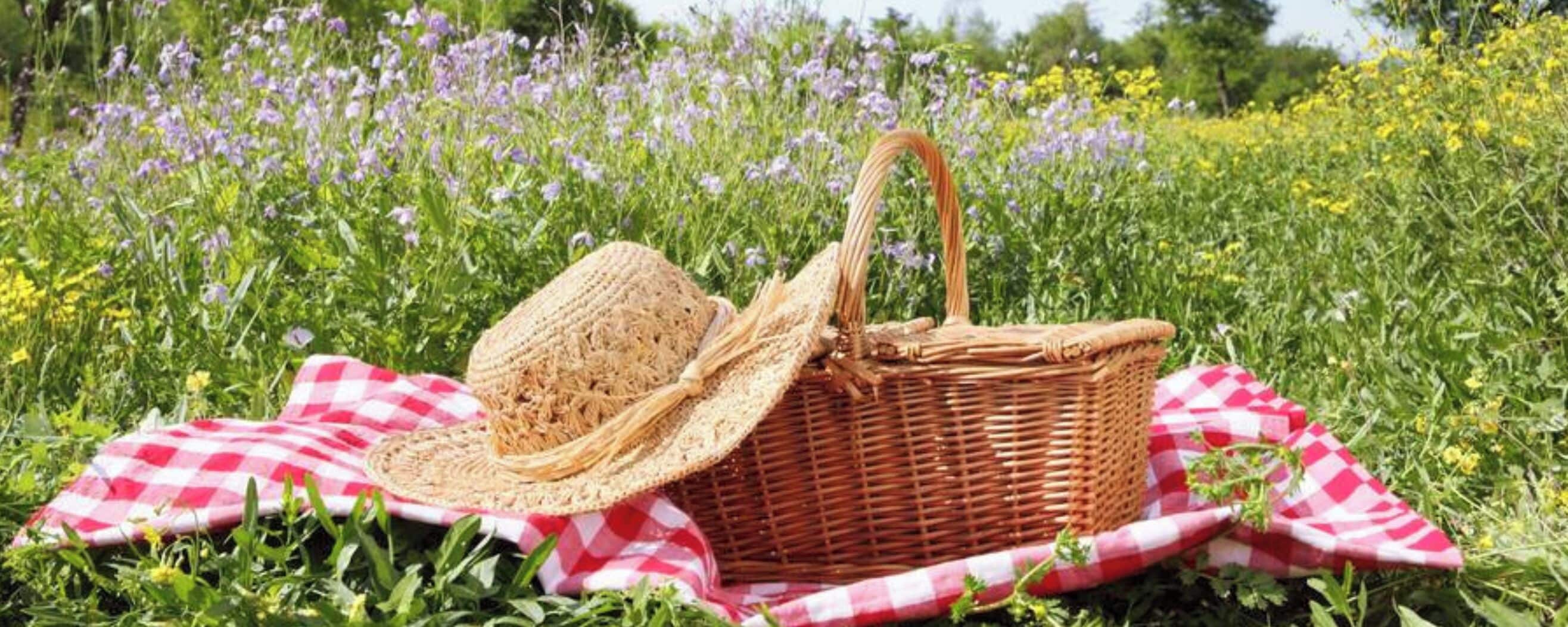 Een rieten picknickmand met handvat en een strohoed, geplaatst op een geruit kleed in een bloemenveld.