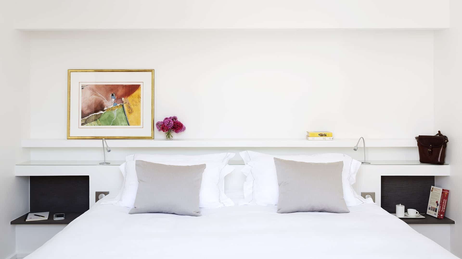 A hotel room with a large bed, two bedside tables, a nightstand, a framed picture, a vase with flowers, a book, and a handbag on the nightstand.