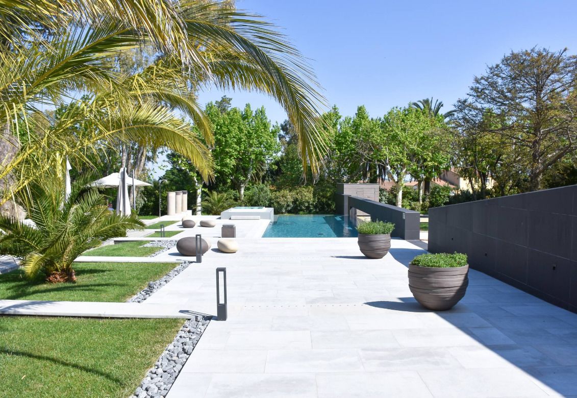 A modern outdoor pool area with a clear view of the pool, surrounded by palm trees, potted plants, and a stone pathway.