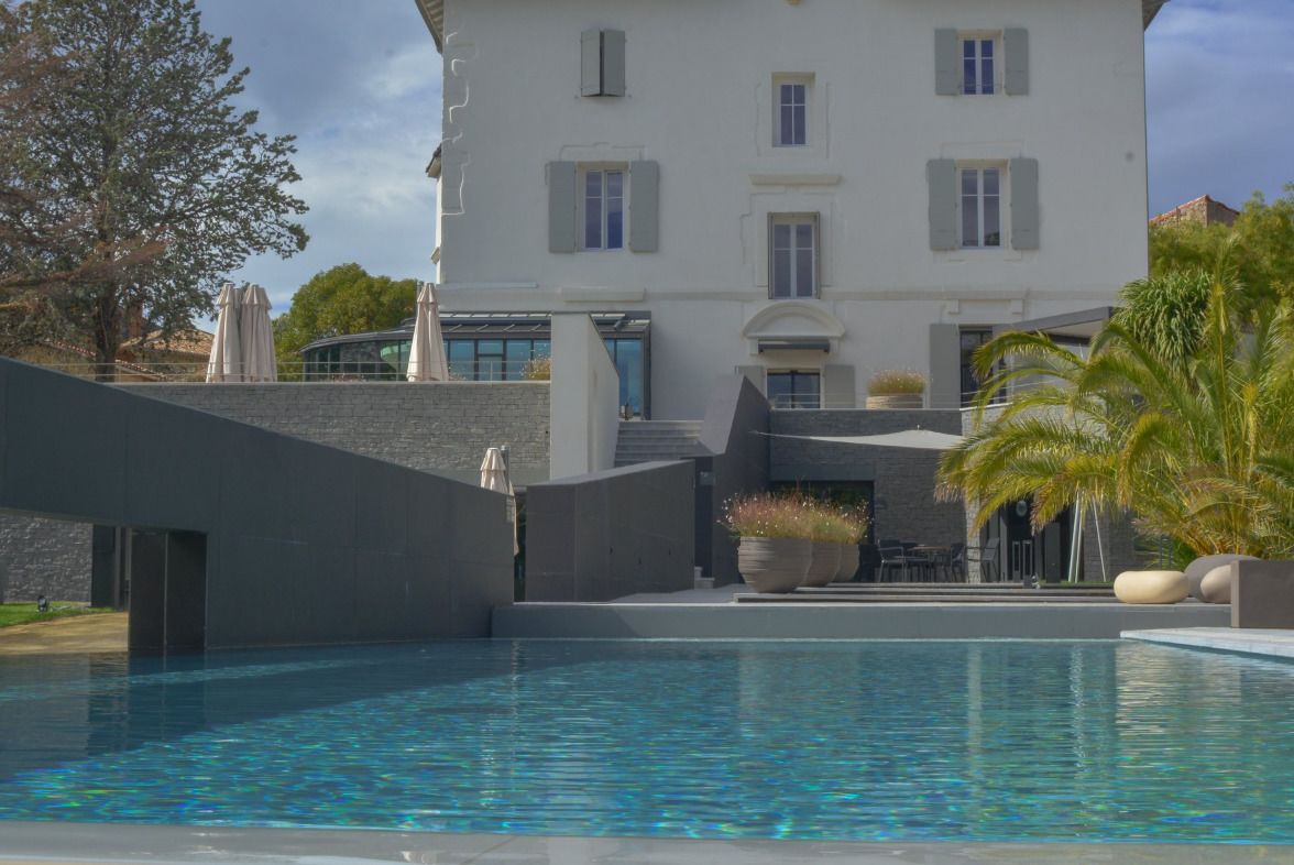 A modern pool area with a large pool, a stone wall, and a building with white walls and shutters in the background.