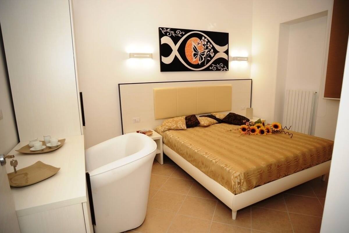 A bedroom with a bed, a nightstand, a white bathtub, and a wall-mounted artwork.