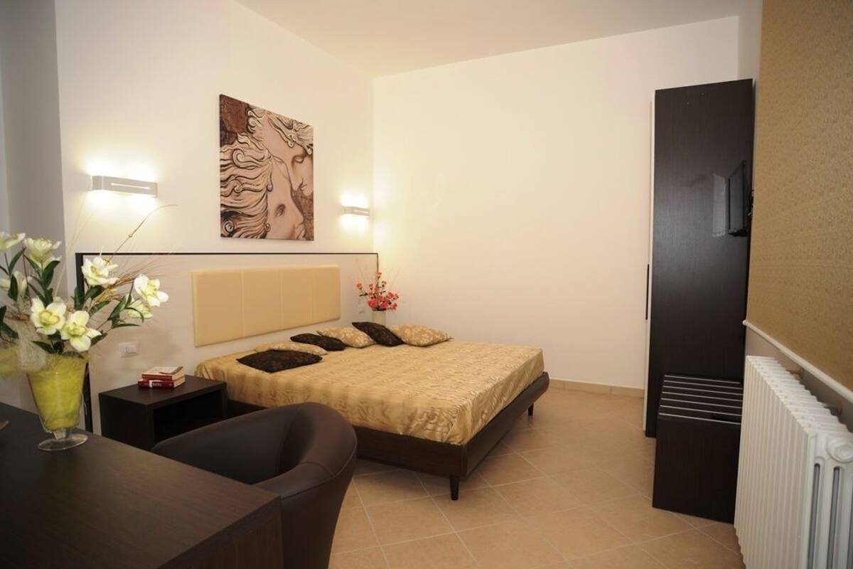 A bedroom with a bed, bedside tables, a desk, a chair, a radiator, and a wall-mounted artwork.