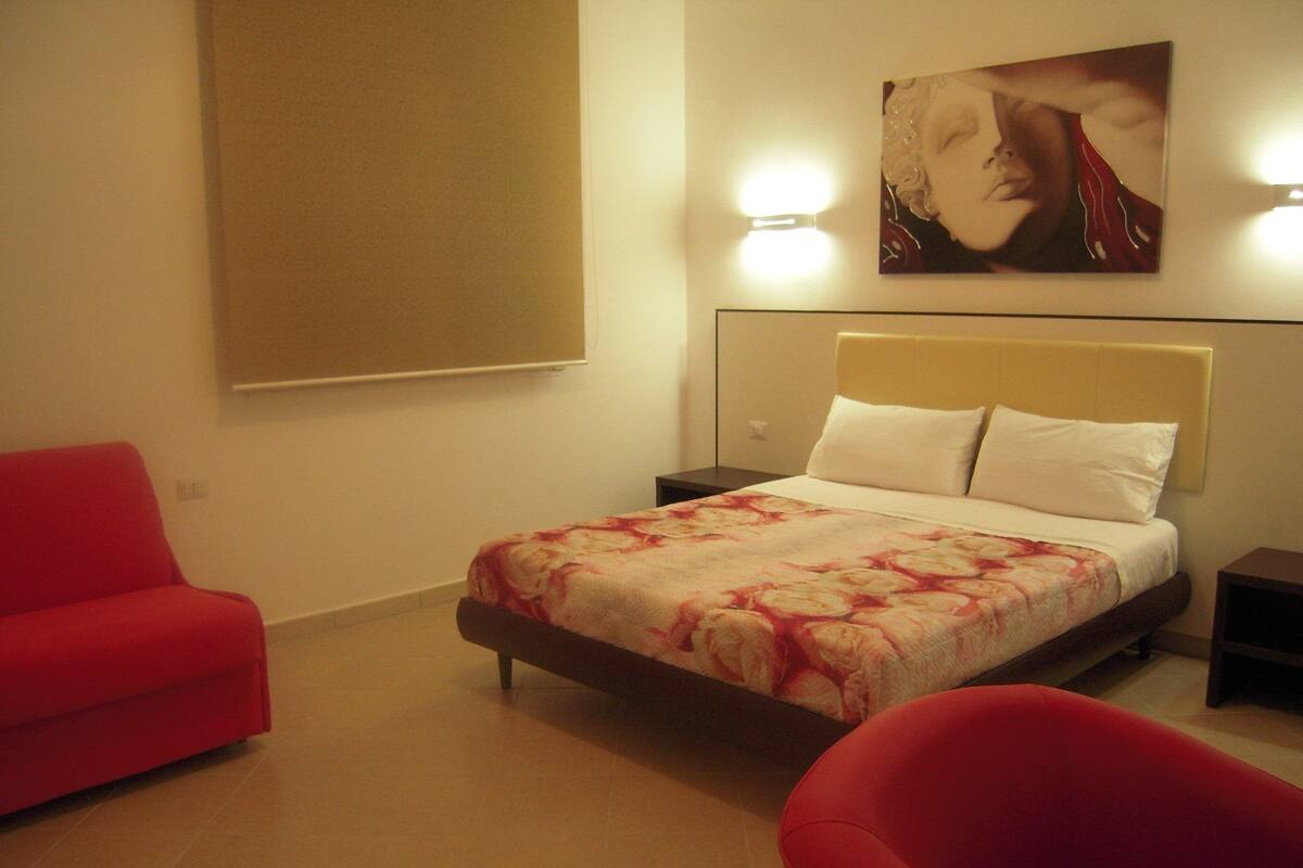 A hotel room with a bed, a red armchair, a nightstand, and a wall-mounted artwork.