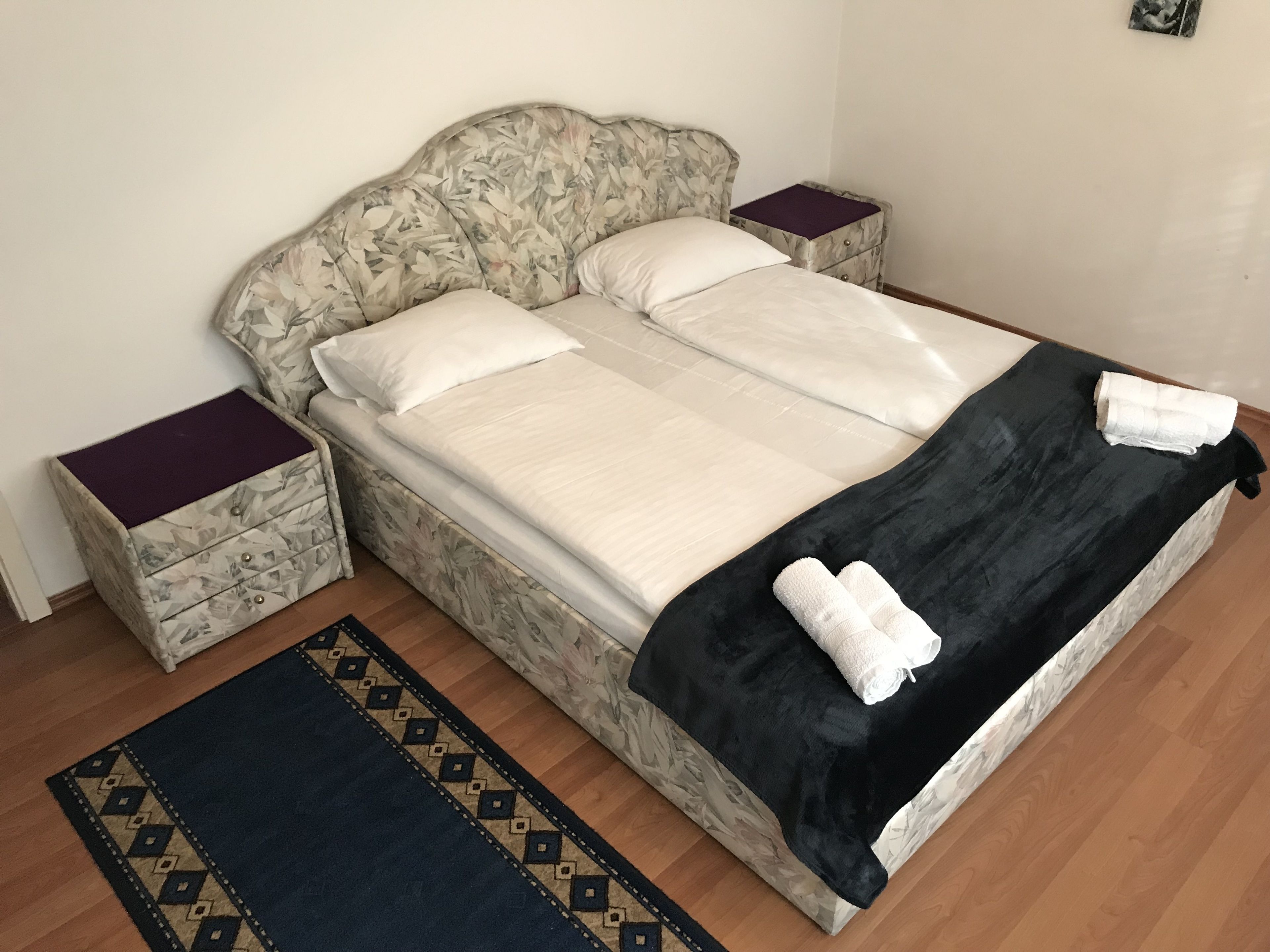 Uma cama com cabeceira florida, duas mesas de cabeceira, um cobertor preto e um tapete estampado.