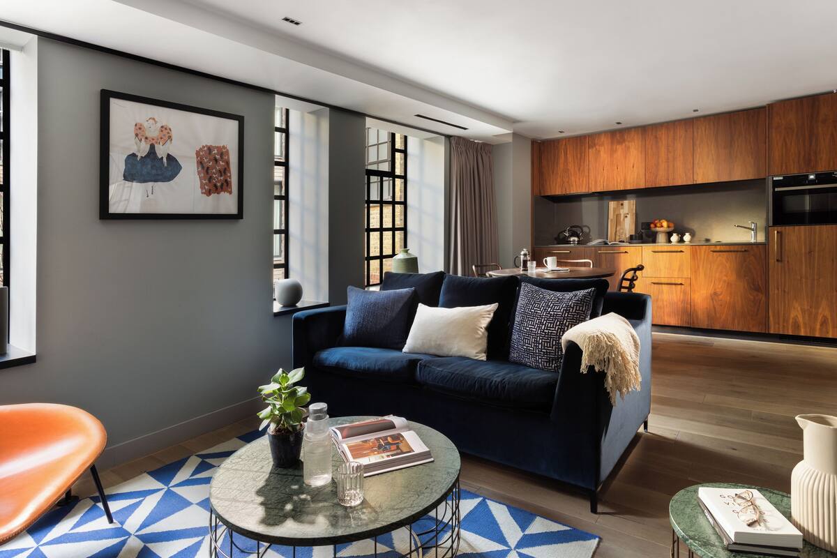 Uma sala de estar moderna com um sofá azul, uma mesa de centro redonda e uma cozinha ao fundo.