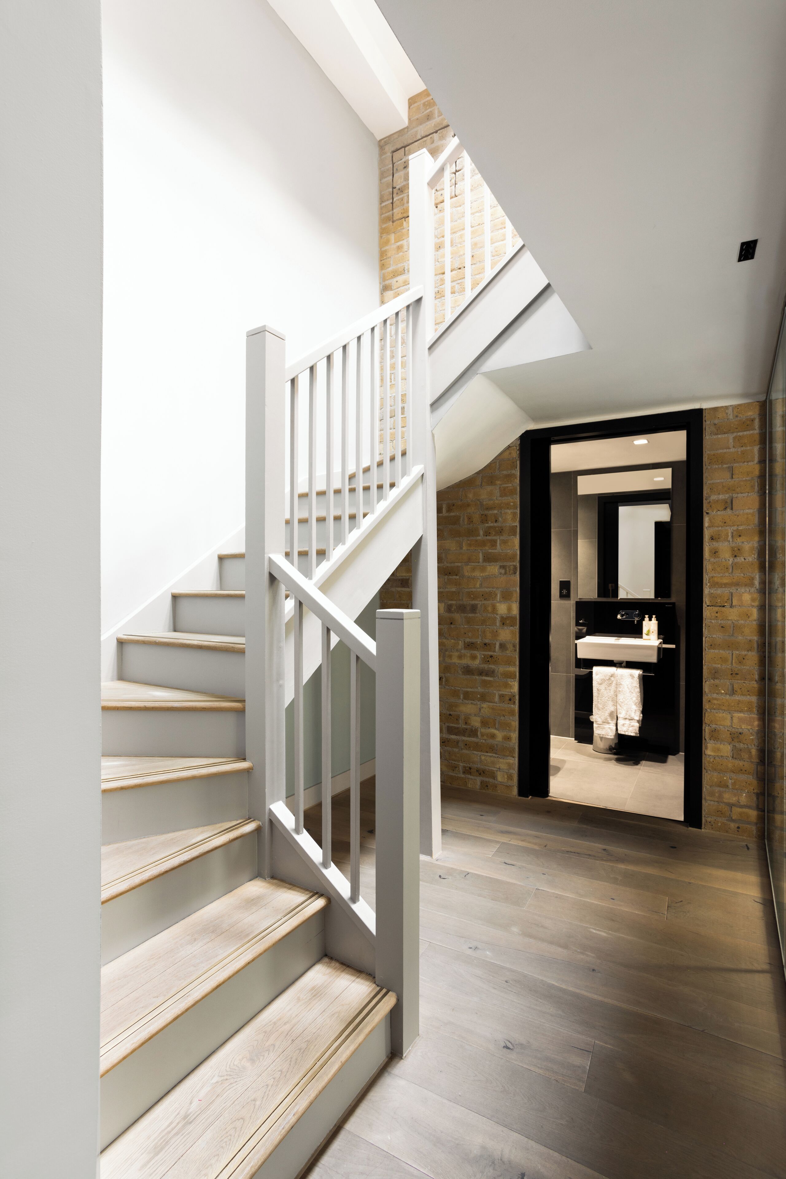 Een moderne trap met houten treden en witte leuningen leidt naar een hoger gelegen gedeelte met zichtbare bakstenen muren en een badkamer met een grote spiegel en wastafel.
