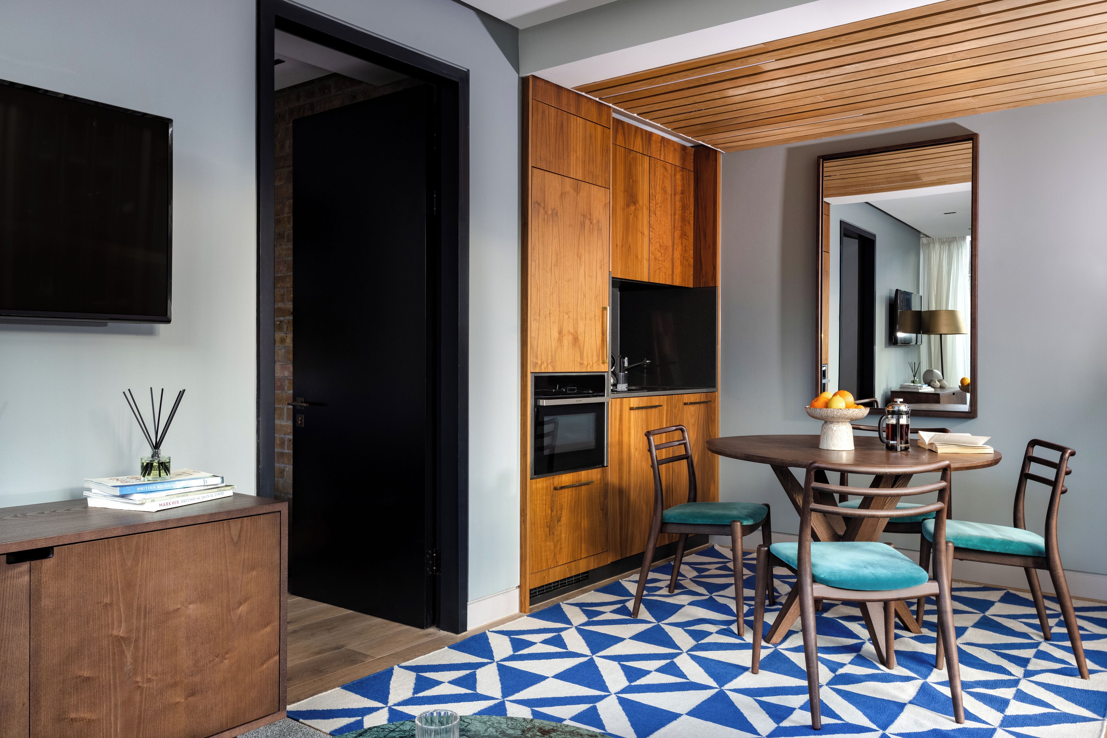 A modern kitchen with wooden cabinets, a black refrigerator, and a dining area with teal chairs and a geometric patterned rug.