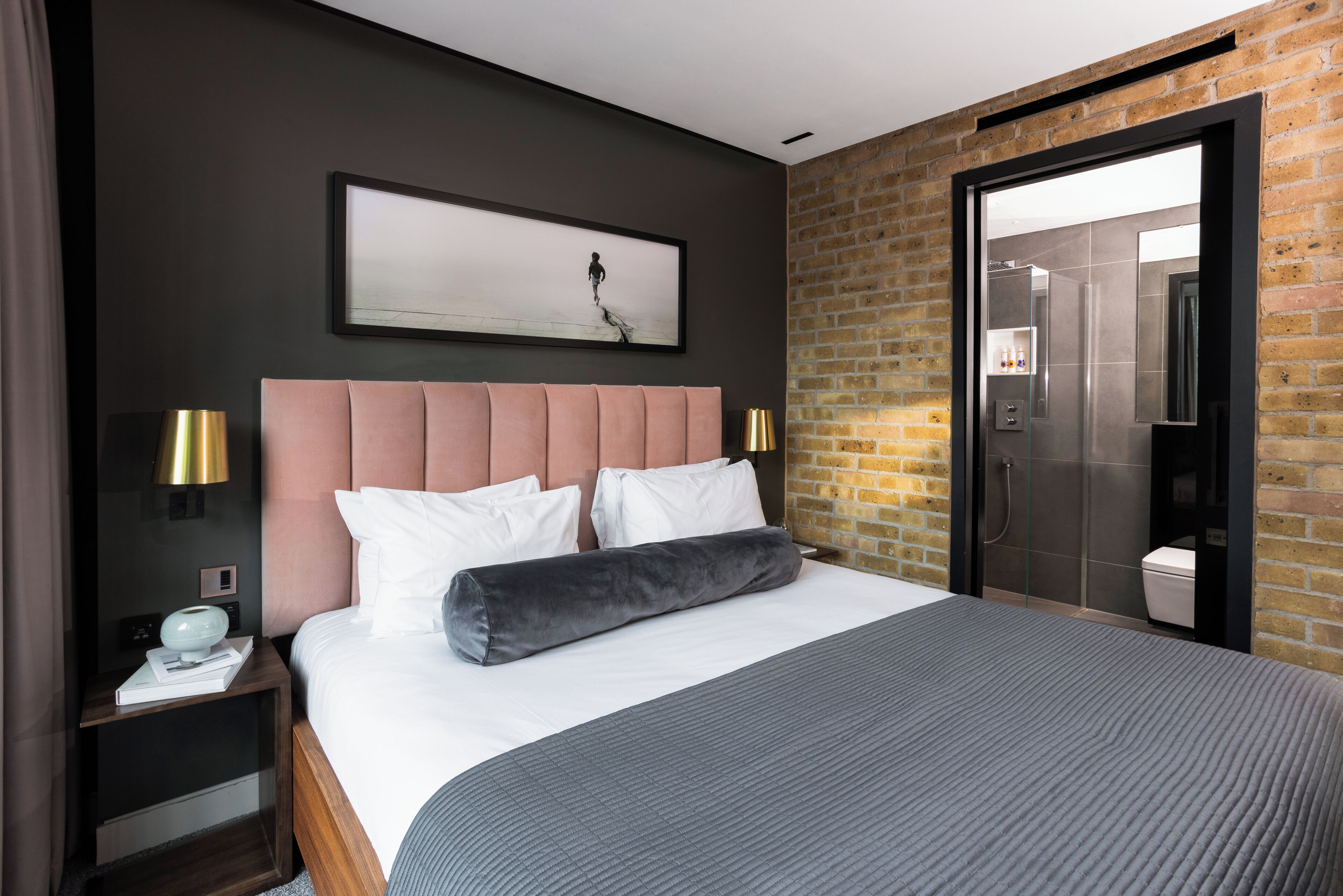 A modern hotel room with a large bed, a headboard, a nightstand with a lamp, a framed picture on the wall, and a brick wall.