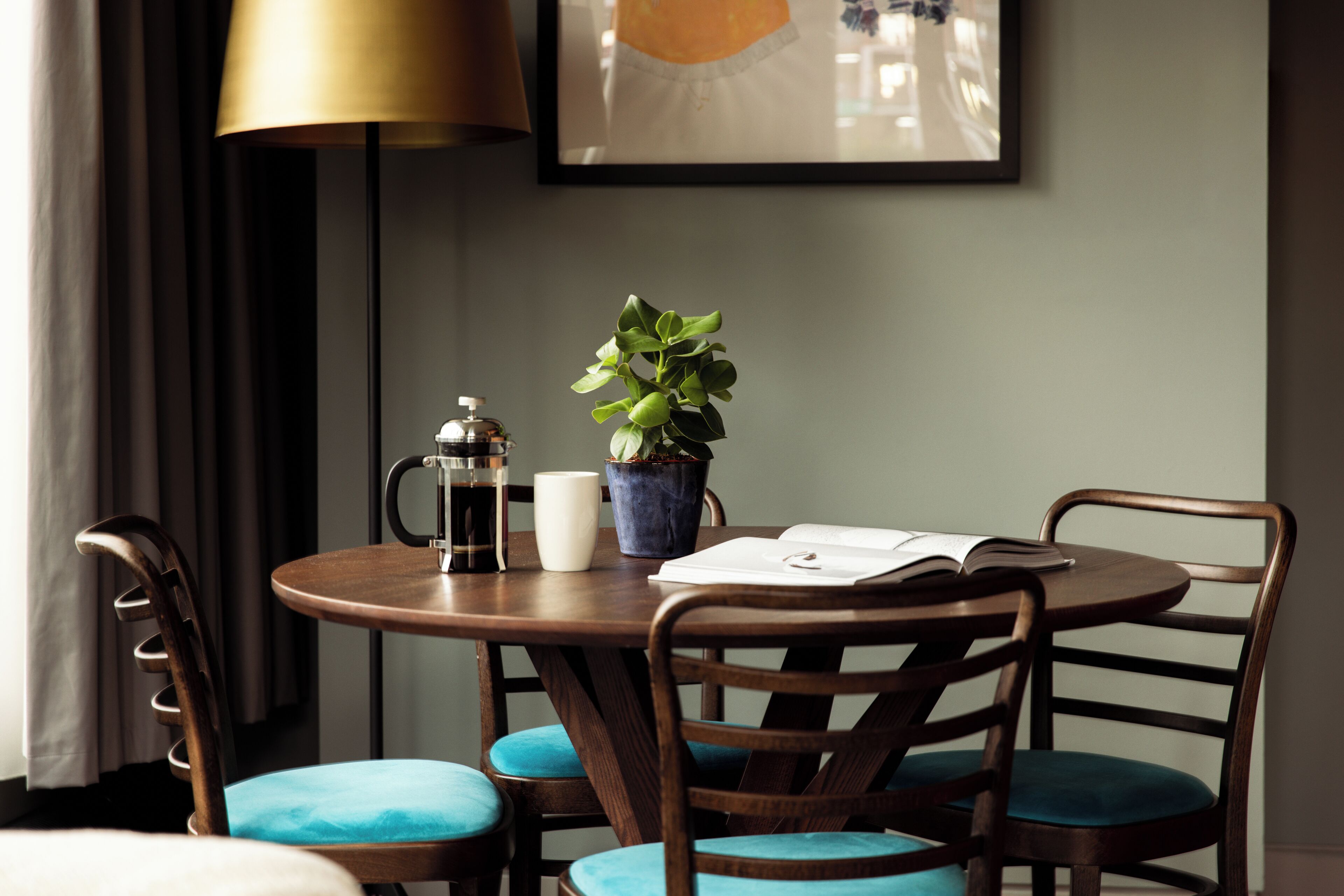 A round wooden table with a coffee pot, a white mug, a potted plant, and an open book. There are four chairs with blue cushions around the table.