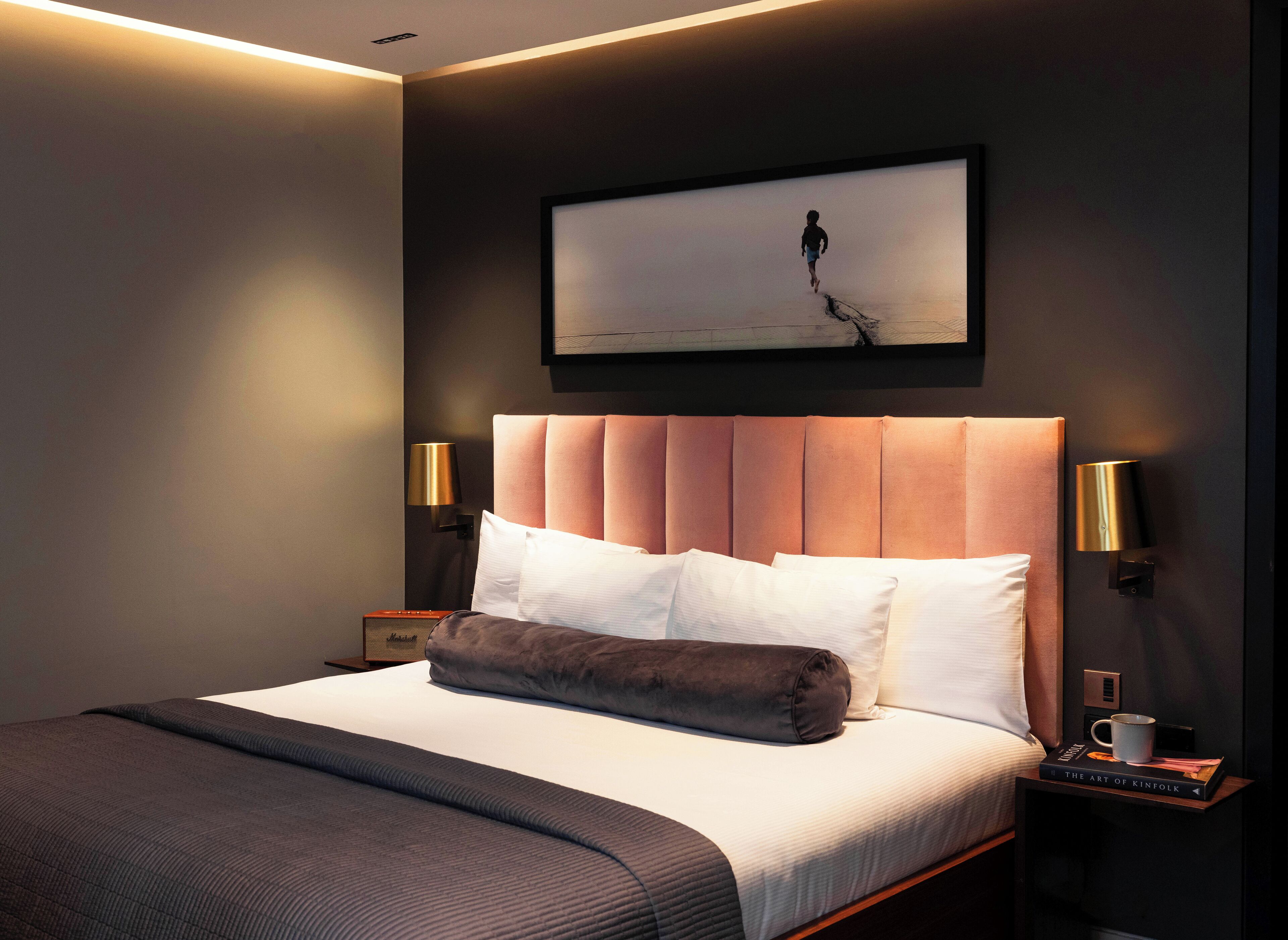 A modern hotel room with a large bed, two bedside lamps, a framed artwork above the bed, and a nightstand with a book and a mug.