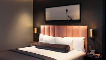 A modern hotel room with a large bed, two bedside lamps, a framed artwork above the bed, and a nightstand with a book and a mug.