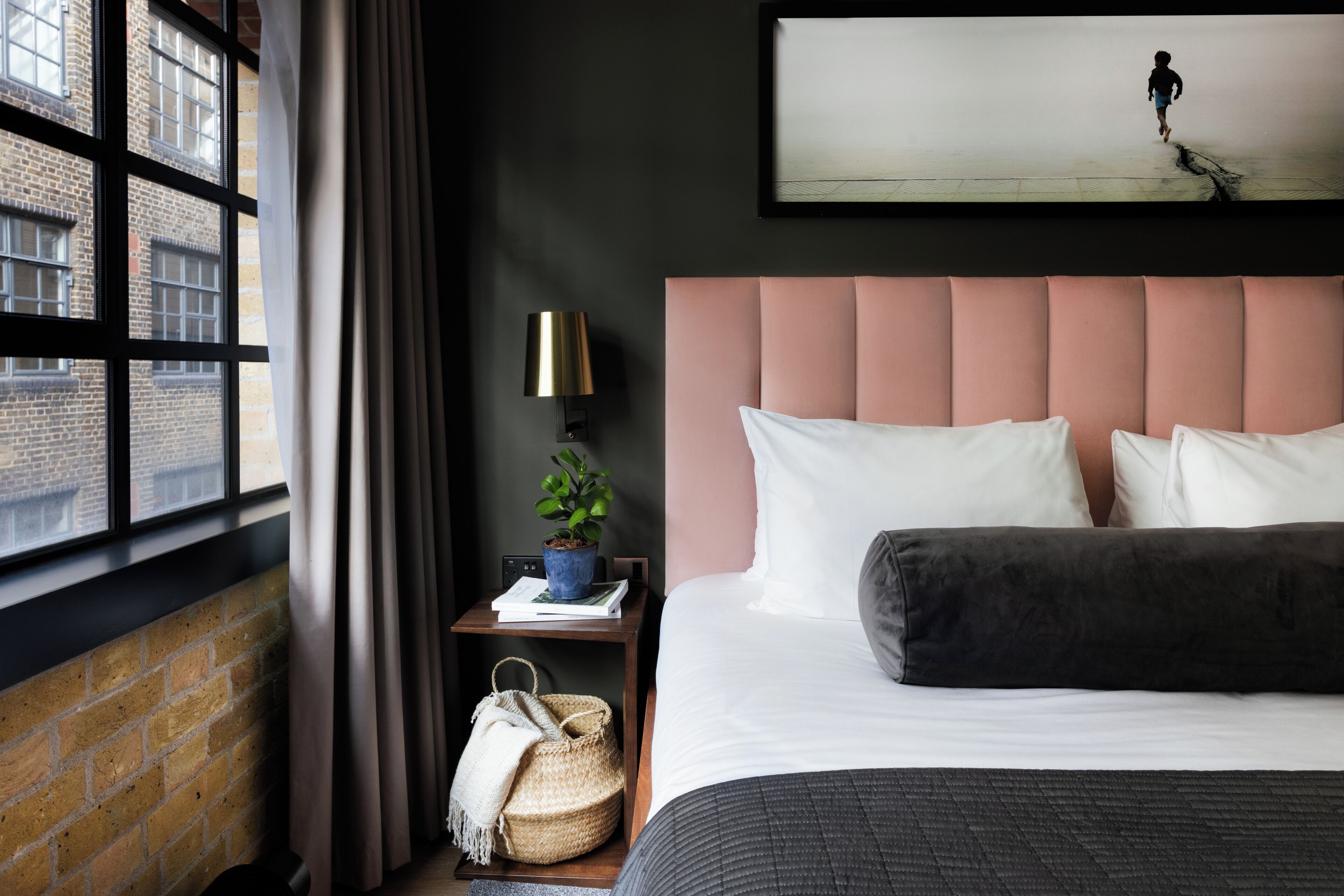 Une chambre avec une grande fenêtre, un lit avec des oreillers blancs et foncés, une table de chevet avec une lampe, une plante et un tableau encadré au mur.