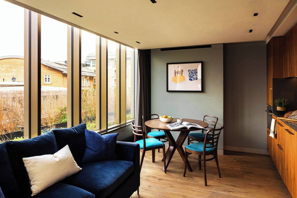 Uma sala de estar moderna com um sofá azul, uma mesa de jantar de madeira e cadeiras. Há uma janela grande com vista para edifícios e árvores.
