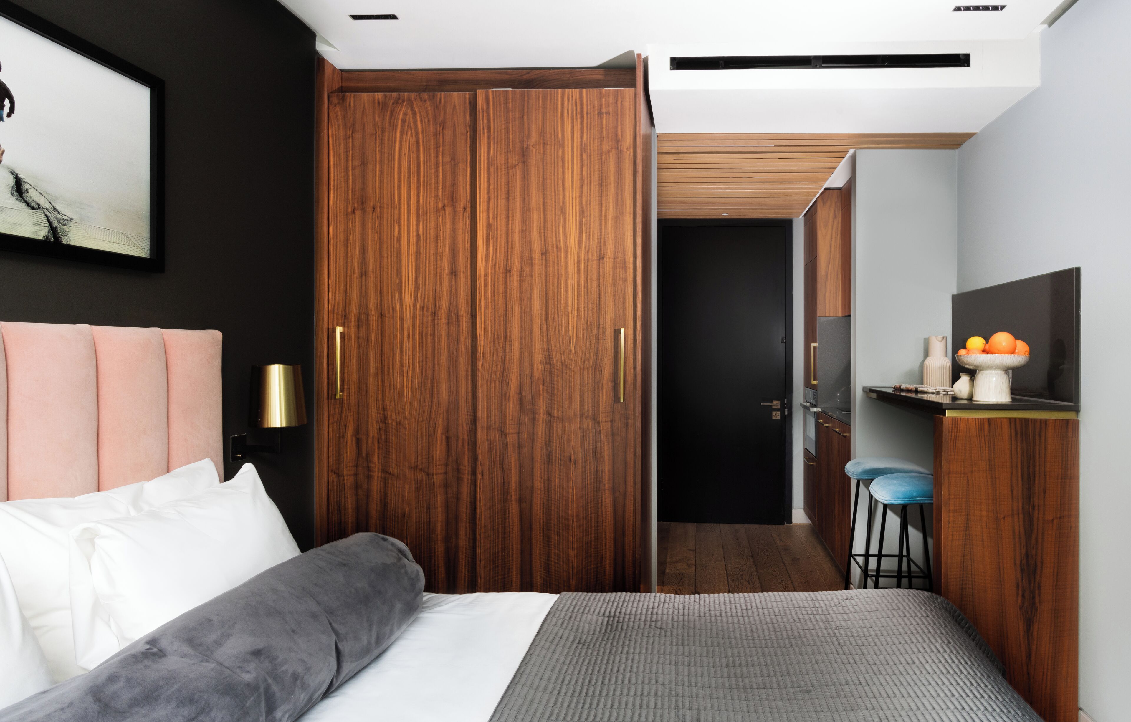 A modern bedroom with a large bed, a wooden wardrobe, a small kitchenette, and a painting on the wall.