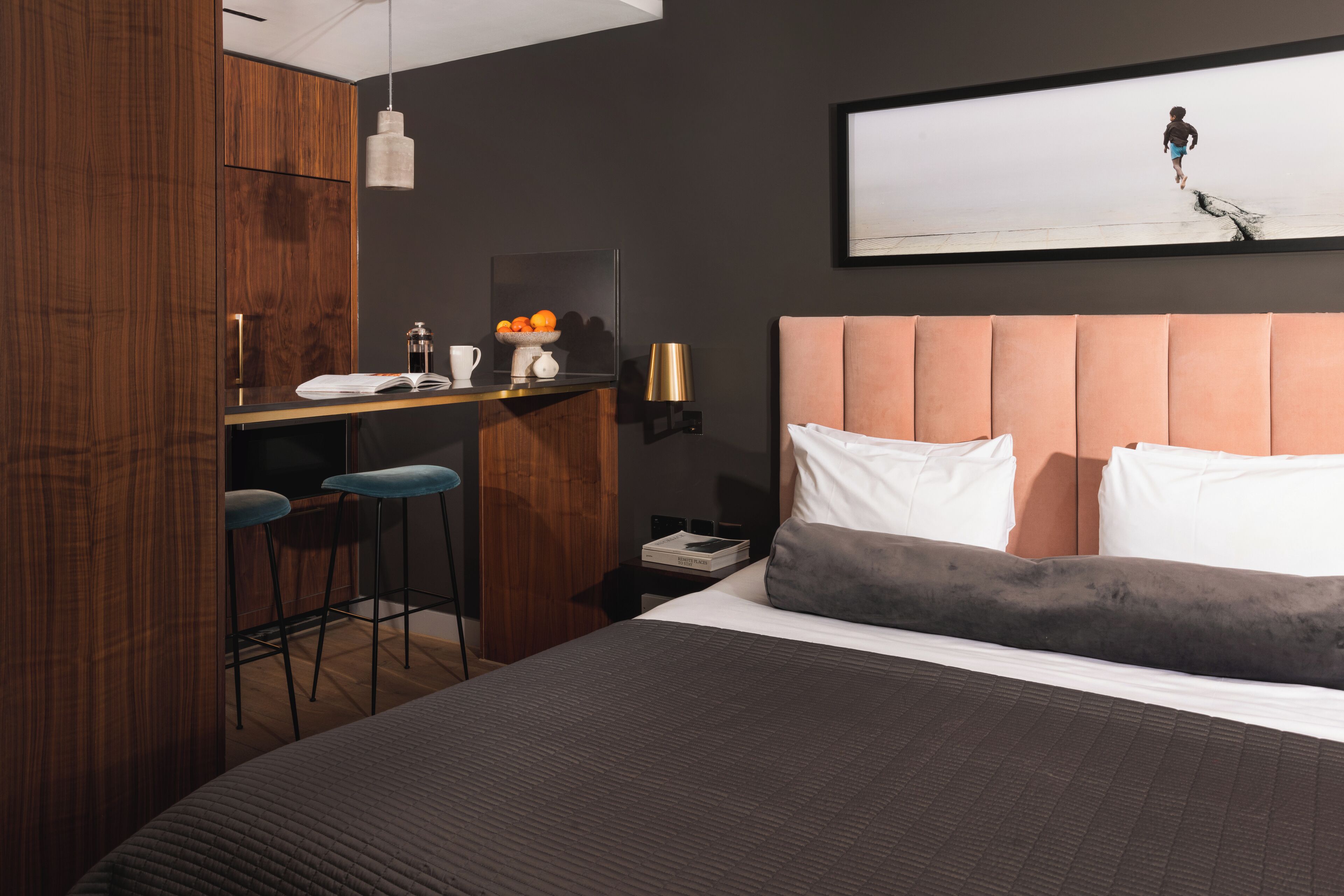 Una habitación de hotel moderna con una cama grande, un escritorio con dos taburetes altos, una mesita con una cafetera y un televisor montado en la pared.