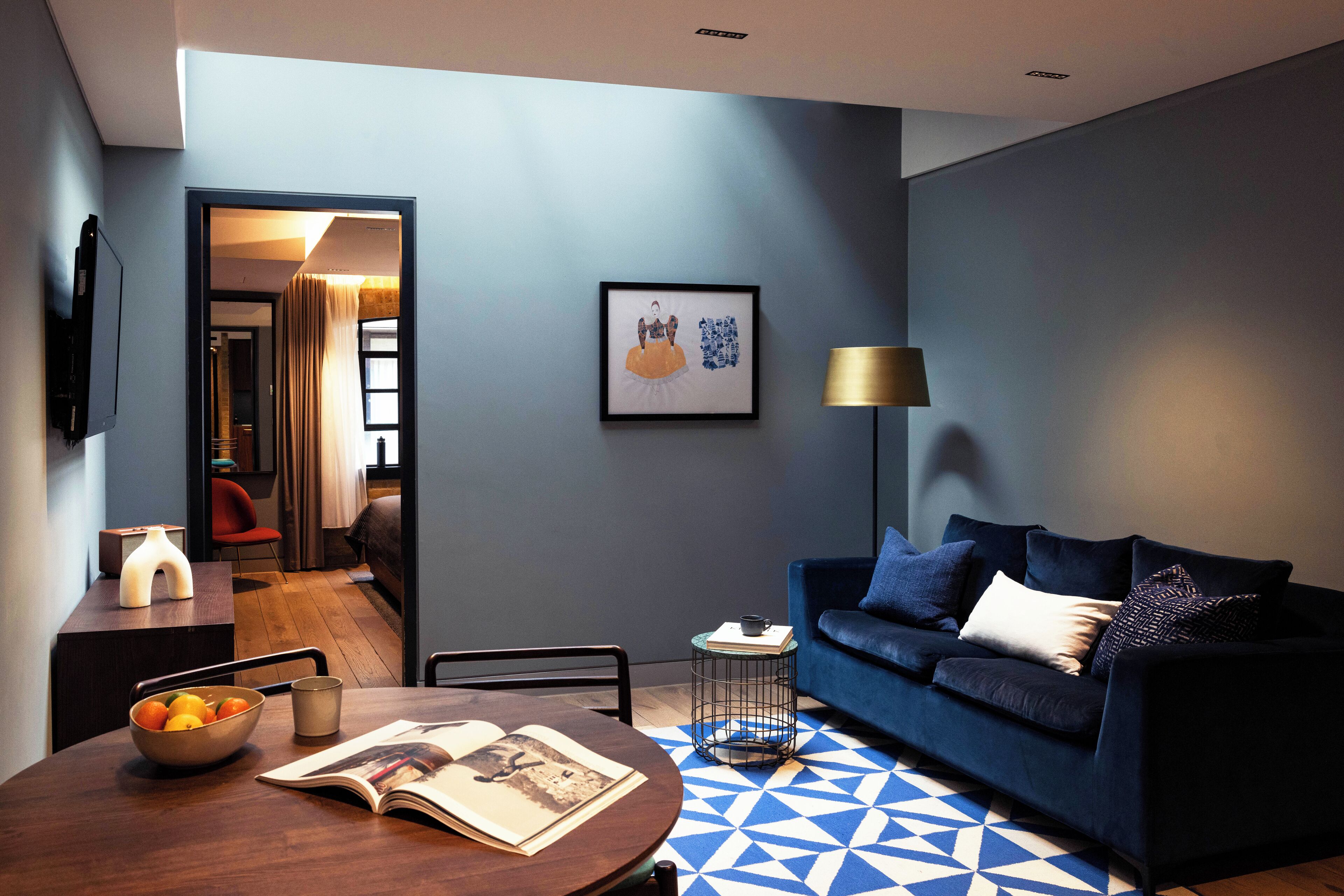 Un salon moderne avec un canapé bleu, une table basse sur laquelle se trouve un saladier rempli de fruits, et un tableau encadré accroché au mur.
