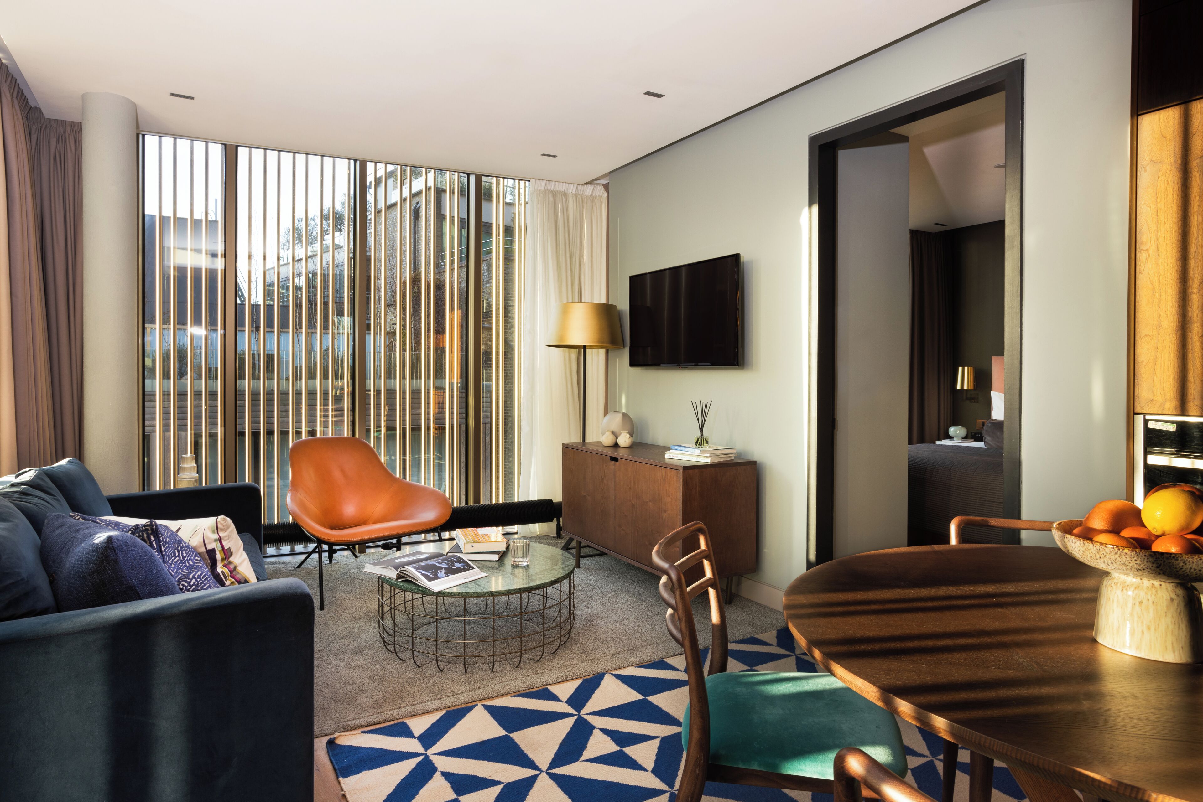 A modern living room with a sofa, a coffee table, a dining table with chairs, a TV mounted on the wall, and a large window with blinds.