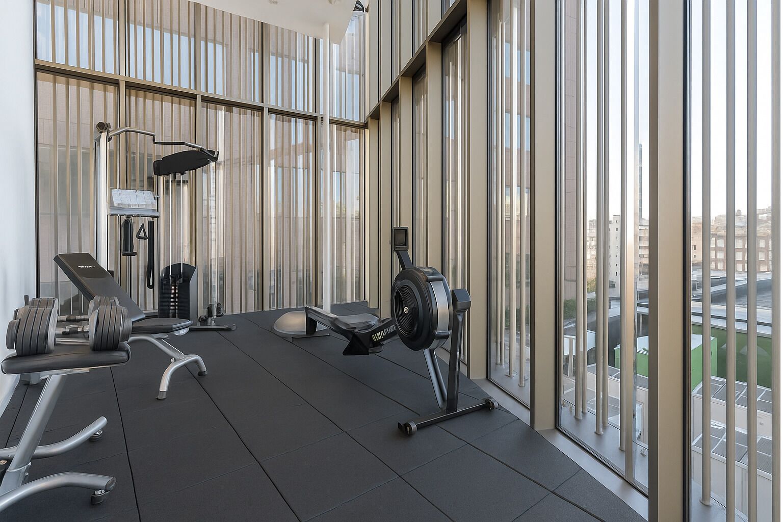 A modern gym with exercise equipment, large windows, and a view of the city.