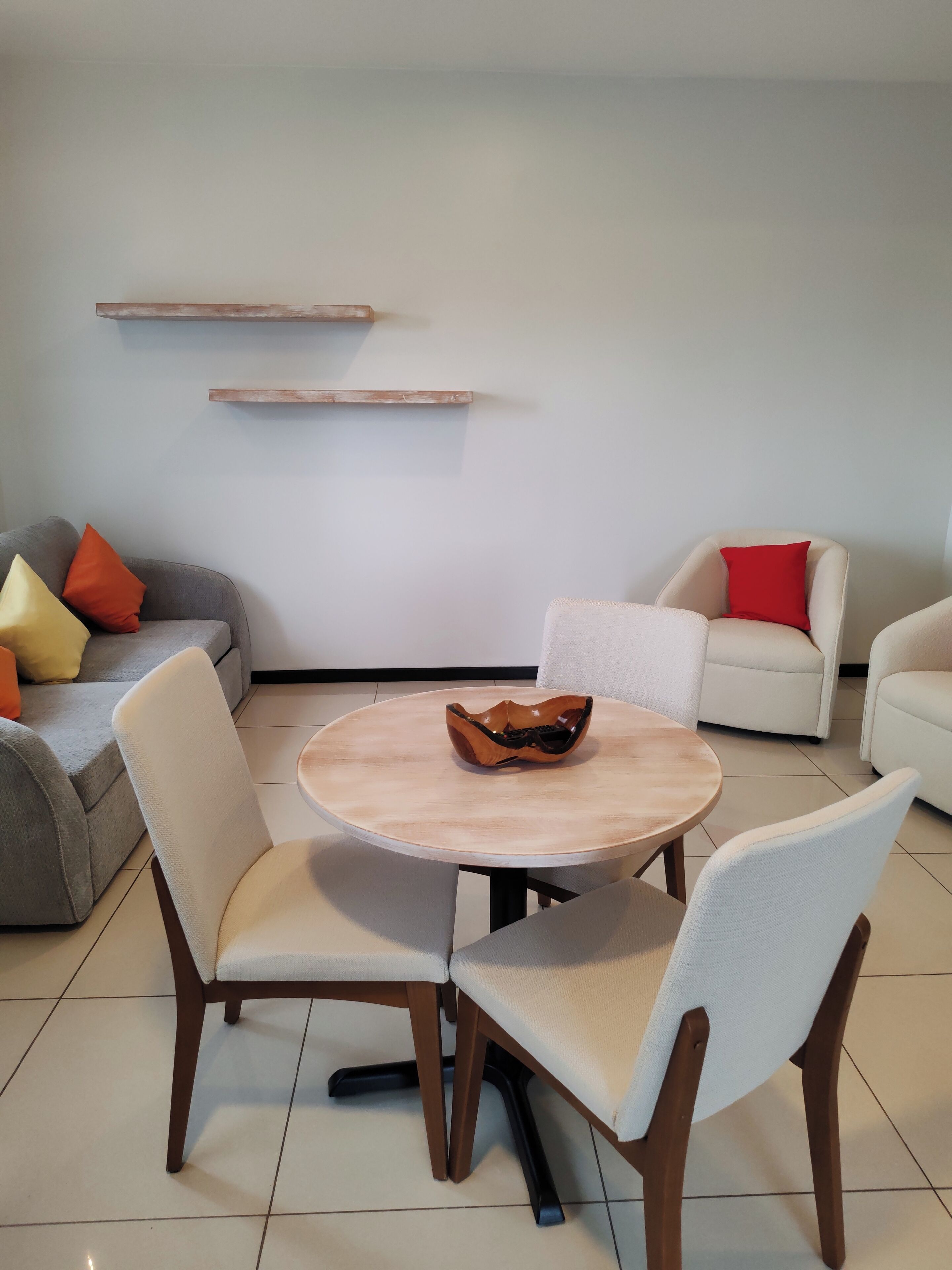 A modern living room with a round wooden table, two white chairs, a grey sofa with orange and yellow cushions, and a white armchair with a red cushion.