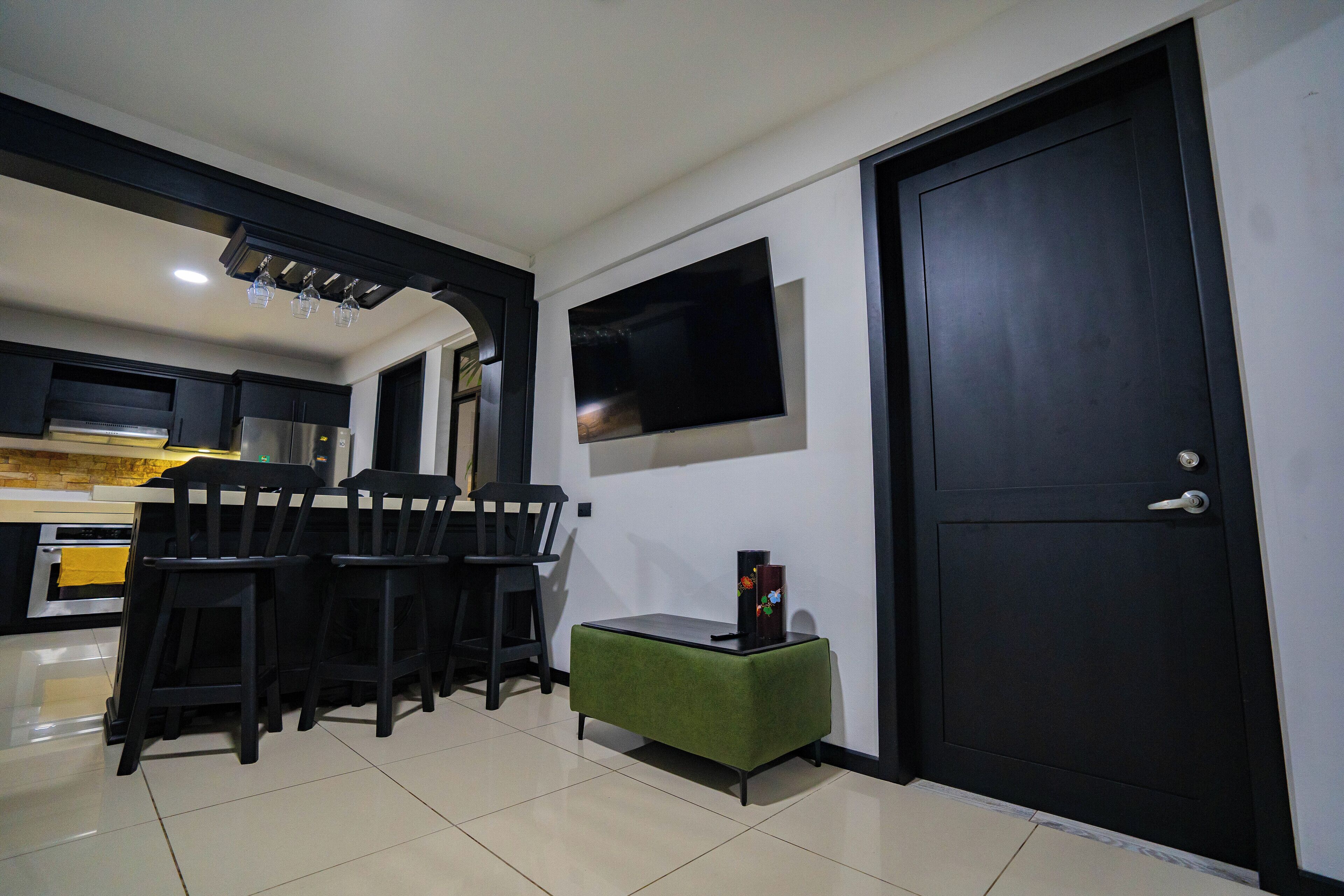 Una cocina moderna con armarios oscuros, una zona de comedor con sillas negras, un otoman verde y un televisor de pantalla plana montado en la pared.
