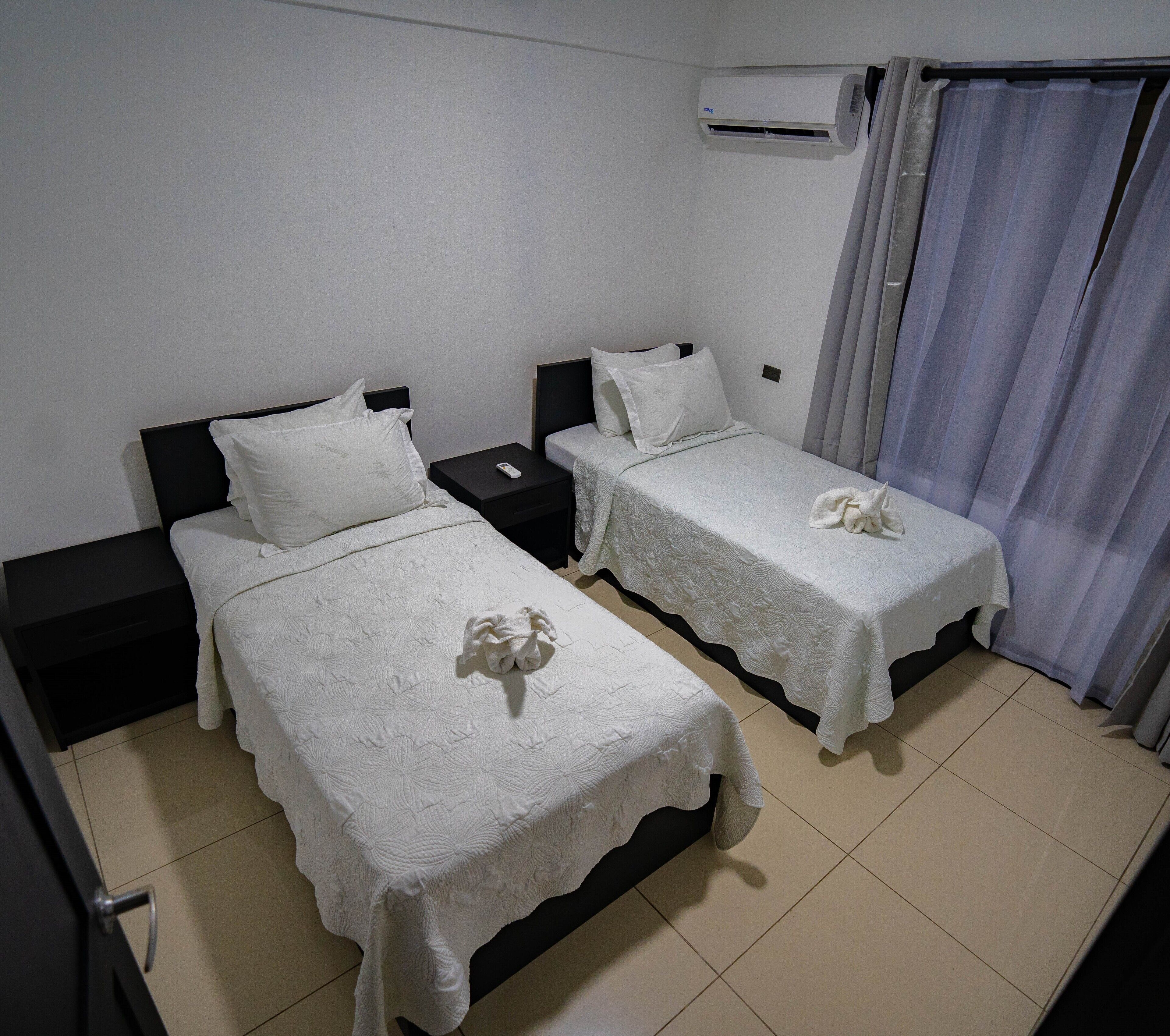 A hotel room with two single beds, each with a white bedspread and a remote control on the nightstand.