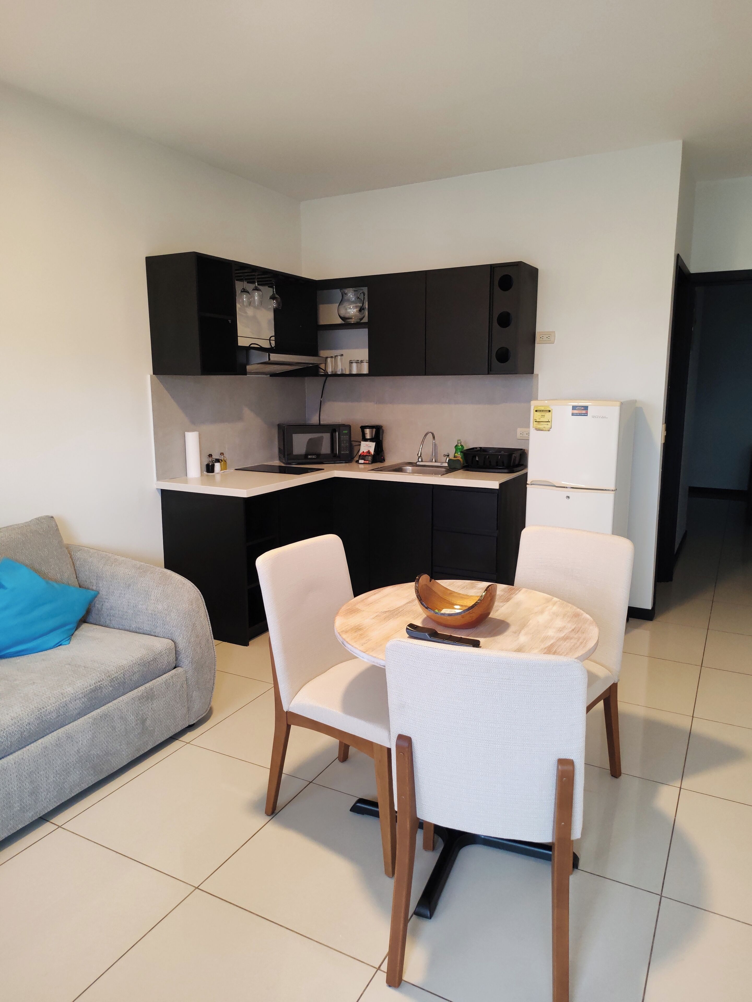 A compact kitchen with modern appliances, a dining table with chairs, and a grey sofa with a blue pillow.