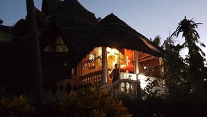 A traditional house with a thatched roof, a balcony, and a person standing on the balcony. - Kivuli Villas (Diani Beach)