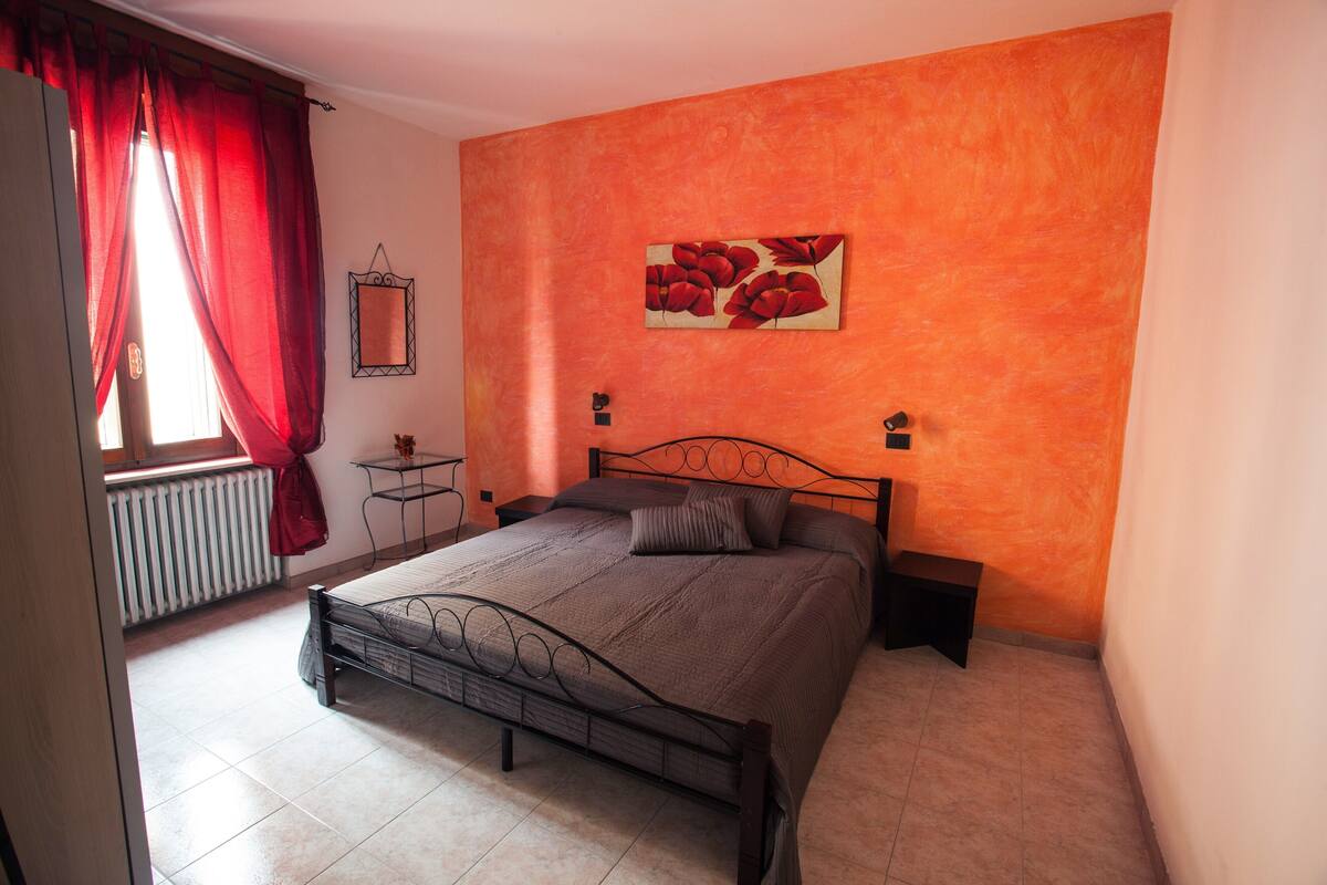 A bedroom with a bed, a window with red curtains, a radiator, and a painting on the wall.