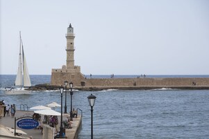 A coastal scene with a lighthouse, a sailboat, and a waterfront promenade with umbrellas and people.