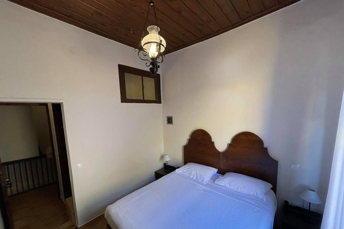 Un dormitorio con techo de madera, cama con ropa de cama blanca, mesita de noche con lámpara y espejo en la pared.