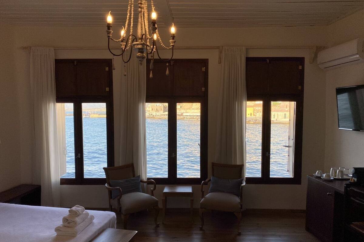 Habitación de hotel con una cama, dos sillas, una mesa y televisor. Amplias ventanas ofrecen vistas al agua y edificios.