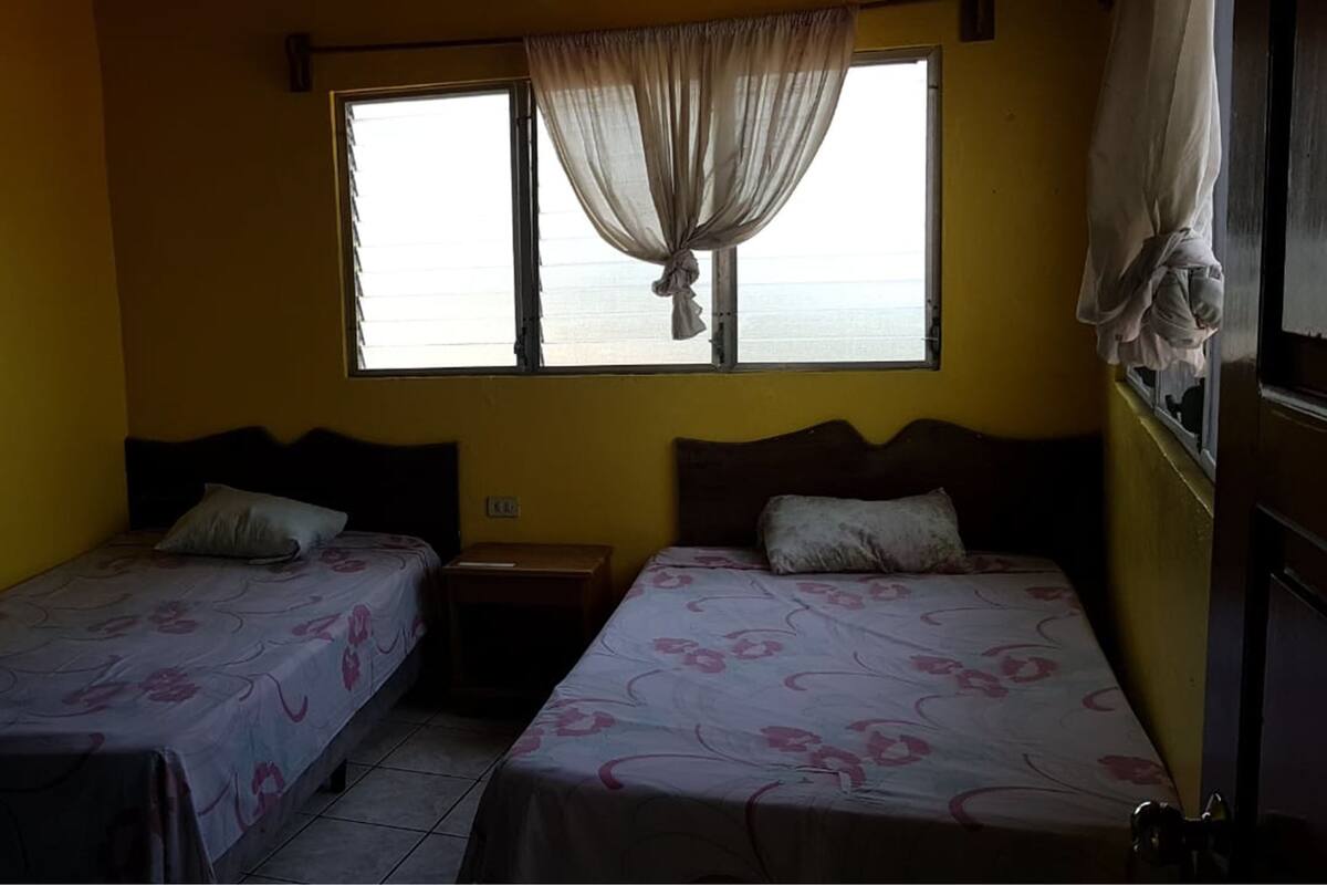 A room with two single beds, a window with curtains, a nightstand, and a view of the sea.