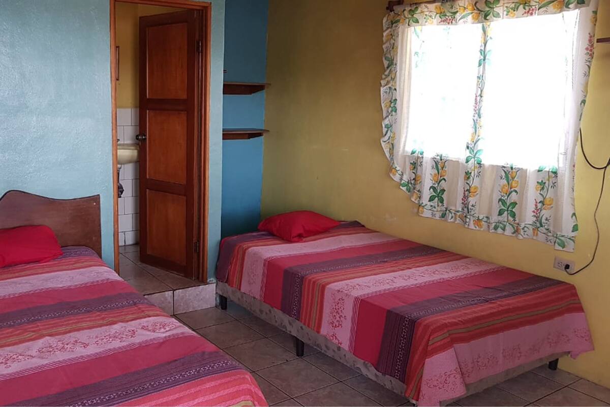 A room with two beds, each with a patterned bedspread and red pillows. There is a wooden door and a window with floral curtains.